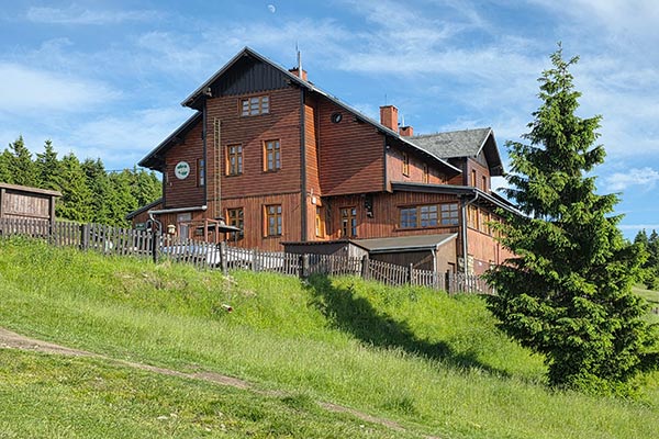 Tradycyjna, kaskadowa bryła drewnianego schroniska turystycznego wtopiona w sudecki krajobraz obok wysokiego świerku